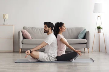 Fotobehang Lotusbloem Covid-19 lockdown, workout at home together, active lifestyle. Millennial calm pacified arab man and woman with closed eyes in lotus position, back to back, practice yoga in living room interior  © Prostock-studio