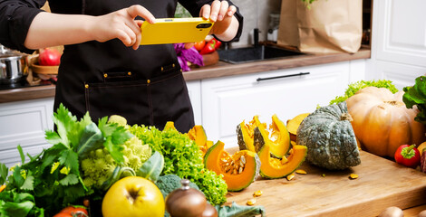 Hands holding smartphone and photographing sliced pumpkin with fresh vegetables on wooden counter...