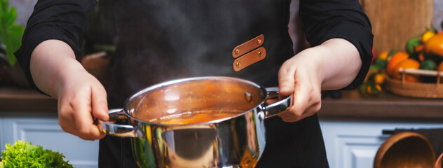 Chef holding steaming pot with pumpkin soup in bright home kitchen, freshly prepared dish among seasonal vegetables for healthy recipe visuals and warm culinary lifestyle content creation.