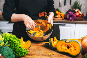 Home cook mixing pumpkin pieces in metal bowl surrounded by fresh green vegetables in bright cozy kitchen. Healthy autumn food preparation, lifestyle cooking and blogging concept