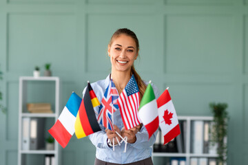 A happy female tutor smiles while holding several diverse flags. She is recommending a foreign language school in her friendly office environment. Her enthusiasm encourages language learning.