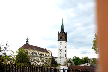 Obraz premium Historic church complex with a tall clock tower rising above tiled rooftops, traditional architecture under cloudy sky, scenic old town landmark framed by trees.