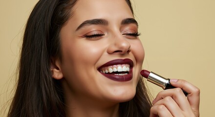 Obraz premium Happy young woman with closed eyes applying dark red lipstick. Close-up beauty portrait of smiling model with professional makeup on beige studio background