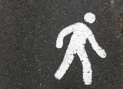 White pedestrian walking symbol painted on dark asphalt road surface. Weathered walking person sign marking designated walk zone for safe street navigation and traffic safety