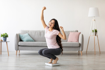 Fototapeta premium Overjoyed Indian woman sitting on scales, gesturing YES, excited over result of her weight loss diet at home. Millennial Asian lady achieving her slimming goal, copy space
