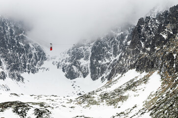 Cable car ascending through misty mountains in High Tatras, Slovakia. Dramatic alpine landscape with snow-covered rocky peaks, glacier valley and fog. Lomnicky stit peak area, winter scenery