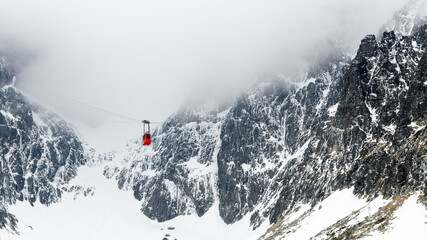 Red cable car gondola suspended over dramatic snowy mountain peaks in misty weather. Steep rocky cliffs covered with snow and ice in High Tatras winter landscape, Slovakia © Arseniy