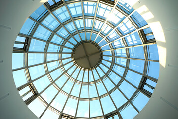 Naklejka premium Circular glass dome viewed from below with radial metal framework, clear blue sky visible through transparent structure, emphasizing symmetry, modern architecture, natural light, and geometric design.