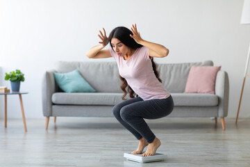Emotional Indian lady terrified of excess weight, sitting on scales in shock at home, copy space....