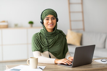 A young Arab female wearing a hijab is focused on her laptop at home. She is using headphones and engaged in her freelance work. Her workspace is cozy and inviting for remote tasks.