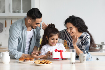 Loving cheerful arabic parents giving birthday girl gift box while having breakfast at kitchen, happy adorable little kid daughter holding festive present, having celebration with mother and father