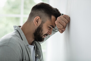 Emotional stress and unhappy, despair, grief and negative emotions, anxiety and agoraphobia. Sad millennial attractive bearded guy resting hand on gray wall and crying, suffer from pain and problems