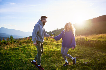 Man and woman hold hands and walk through sunlit meadow. Sun sets behind, casting warm glow over scene. Their laughter and flowing landscape capture moment of happiness and connection.