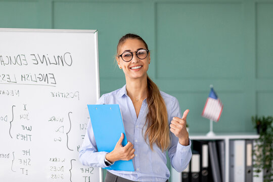 Smiling teacher stands in front of a whiteboard while giving English lessons. She holds a clipboard and shows a thumbs up, expressing satisfaction with the language school and its services.