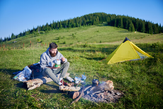 Bearded man eating from food pouch. Tourist in gray hoodie and green pants sits on log near campfire. Beside him, camping gear and yellow tent add to scenic outdoor setting.