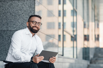 Business news. Happy mature businessman using digital tablet sitting outdoors near modern office center in urban area cityscape, looking aside and smiling, free space