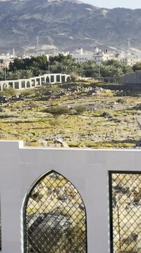 Sacred Shuhada Badr burial ground in a serene setting. View features rocky mountains overlooking Madinah outskirts, green vegetation, stone terrain, traditional walls, and distant urban buildings.