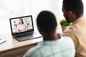Fototapeta premium Back View Of Black Couple Having Video Call Online Consultation With Professional Manager Sitting At Laptop Consulting Distantly At Home. Modern Communication. Selective Focus