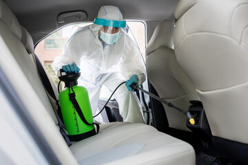 Man in protective hazmat suit with mask disinfecting inside car, wipe clean surfaces that are...