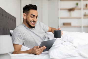 A happy young Arab man relaxes in bed, sipping coffee from a cup. He smiles while using a digital tablet, reading the news or browsing the internet, enjoying a peaceful morning at home.