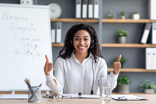 Online teacher, distant learning language, video call, recording vlog, covid-19. Smiling millennial black female tutor in headphones on blackboard background, shows thumbs up, looks at webcam at home