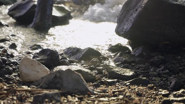 Lake water shines in the sun as sea washes ashore on rocks and pebbles on beach. Sunset or sunrise with warm orange color. Peaceful and clam water reaches the coast. Lens flare from sunlight on spray