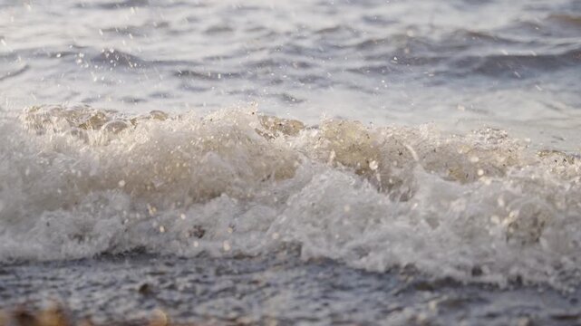 Waves crash gently onto sandy shore. Sunlight glints off foamy water. Ripples roll toward beach in slow motion. Soothing nature scene, ideal for calm visuals. No people, animals, or plants visible