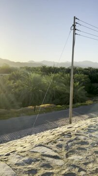 A rural oasis with palm trees, small village buildings, distant mountains under warm golden-hour light, highlighting the solemn Maqbarat Shuhada Badr sign at the historic Islamic battlefield site.