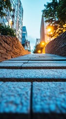 Low-angle view of a stone pathway leading upwards towards city buildings at dusk, with warm streetlights illuminating the scene and trees providing a natural contrast to the urban environment.