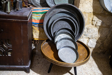 A stack of vintage copper trays resting on a small wooden table next to an antique cabinet in an old stone room in Nazareth.