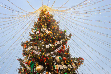 Moscow, Russia, 29 December 2025 A low angle view of a Christmas tree adorned with ornaments and postcards, with strings of lights radiating outwards from the top against a pale blue evening sky.