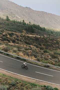 A lone cyclist rides a black road bike along a winding mountain road, surrounded by rugged hillsides and sparse pine trees. Emphasizes endurance, outdoor activity, and scenic travel.