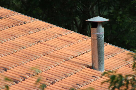 Close-up of a terracotta tiled roof with a metal chimney pipe and lush green foliage in the background