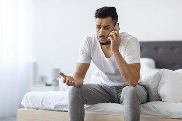 A young Arab man is sitting on his bed in a bedroom, visibly worried while engaged in a difficult phone conversation. His expression shows frustration and concern as he talks.