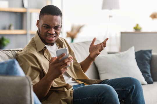 Problem With Phone. Displeased Black Man Using Smartphone Having Issue With Connection Or Mobile App Sitting On Sofa At Home. Disconteted Cellphone User Reading Negative Message Or News