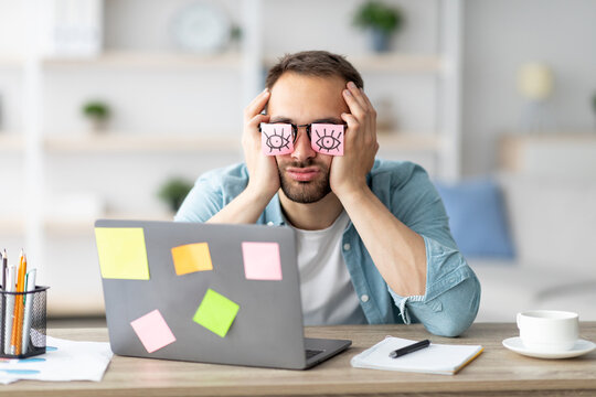 Tired young man having stickers with open eyes drawing on his glasses, sleeping in front of laptop at home office. Exhausted millennial man overworking, napping, failing to meet deadline