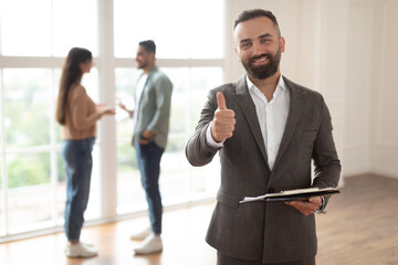 Successful Deal, House Ownership Concept. Portrait Of Smiling Professional Real Estate Agent In Suit Showing Thumb Up Sign Like Gesture, Selective Focus. Buyers Couple Talking In Blurred Background
