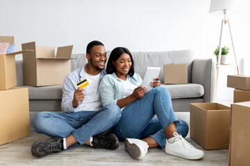 Excited black couple choosing furniture online, buying goods for their new flat, using digital tablet and credit card, sitting on floor among unpacked boxes and shopping in internet