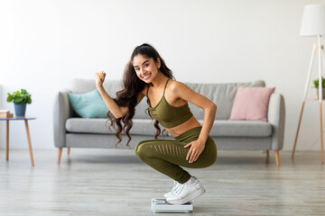 Triumphant young Indian woman sitting on scales, making YES gesture, happy with result of her...
