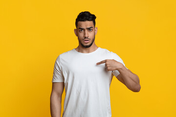Confused handsome bearded young muslim guy in white t-shirt pointing at himself, having puzzled...
