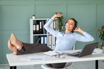 A female entrepreneur takes a break at work, sitting comfortably in an armchair with her feet on the table, enjoying music through headphones and smiling. She appreciates her favorite songs.