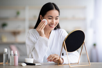 Beautiful asian woman cleaning her face, using cotton pads and cleansing product, looking at mirror in bedroom. Young attractive korean lady using face toner and cotton pad, home interior, empty space © Prostock-studio