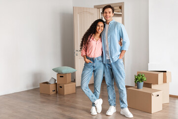 Young couple stands cheerfully in their new apartment surrounded by cardboard boxes after moving in. They embrace each other, showing their happiness for home ownership and new beginnings.