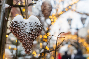 Naklejka premium Close up of heart shaped pine cone ornament covered with snow hanging on branch with Christmas lights, Moscow, Russia.