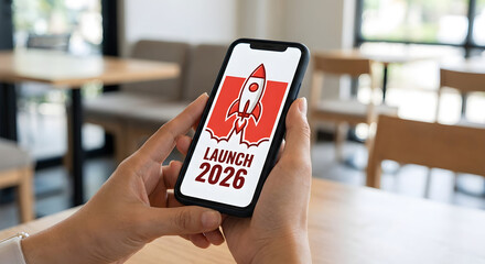 Launch 2026 business startup concept on smartphone screen, hands holding mobile phone with rocket icon, new year goals, app release, digital marketing strategy, future growth planning, blurred cafe.