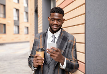 Happy african american entrepreneur holding takeaway coffee and using smartphone while walking outdoors during break and spending time near office center