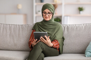 Senior Muslim Female Using Digital Tablet Reading Book Online Sitting On Couch At Home. Modern...