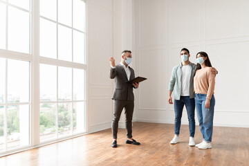 Fototapeta premium Meeting With Realtor During Pandemic. Professional Real Estate Agent In Suit And Face Mask Showing Modern House With Floor-to-Ceiling Windows To Beautiful Young Couple Purchasing Or Renting New Home