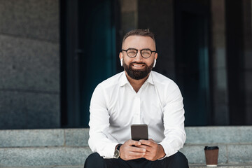 Confident businessman with smartphone in his hands wearing earphones and listening music or podcast outdoors, sitting on stairs near office. Mature man enjoying break and drinking coffee