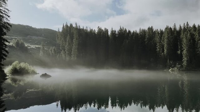 Survol en drone d'un lac de montagne paisible avec brume matinale et reflets de for&ecirc;t. 